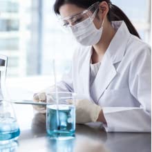 Woman wearing lab coat, face mask, gloves, and protective glasses in a lab with fluid in a beaker.